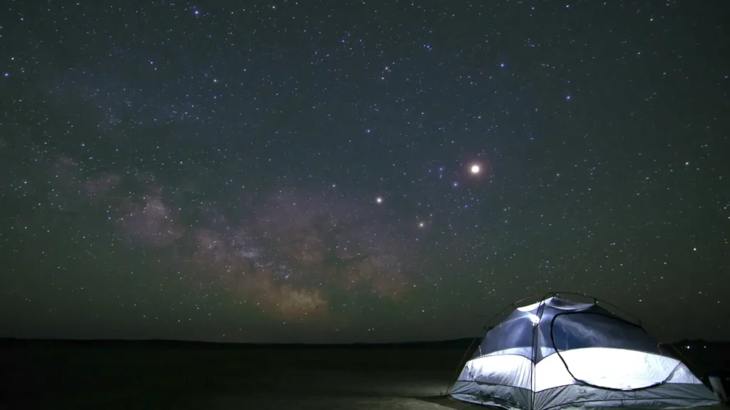 Illuminated tent under a star-filled night sky with a clear view of the Milky Way, set in a remote and open desert landscape.