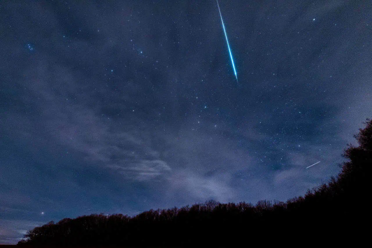 A bright meteor streaks across a starry night sky above a forest silhouette, with clouds scattered and constellations visible in the background.